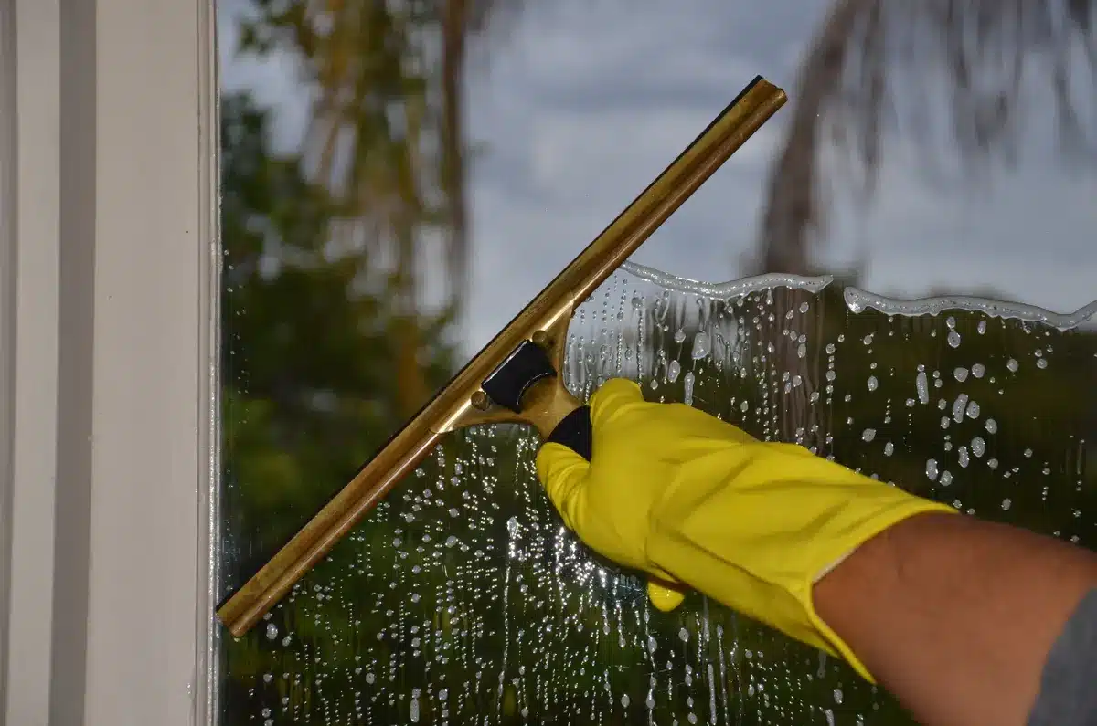 Hand mit gelbem Handschuh zieht Fensterabzieher über nasse Kunststofffenster-Scheibe ab