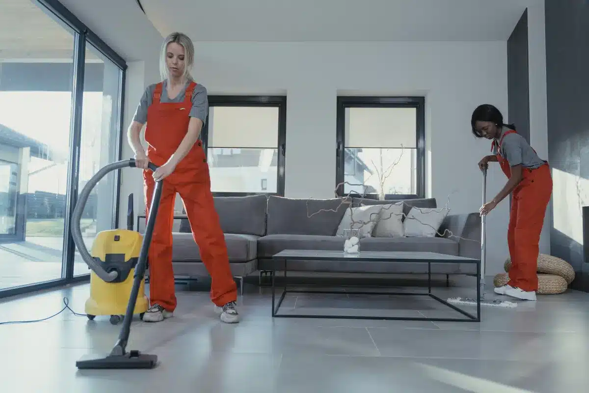 Professional cleaners in red uniforms vacuuming and cleaning a modern bright living room with sofa and windows