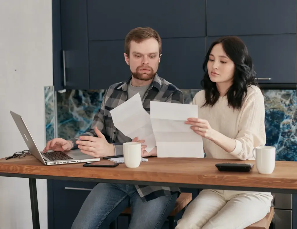 Couple reviewing invoices and documents at wooden kitchen table with laptop and coffee cups
