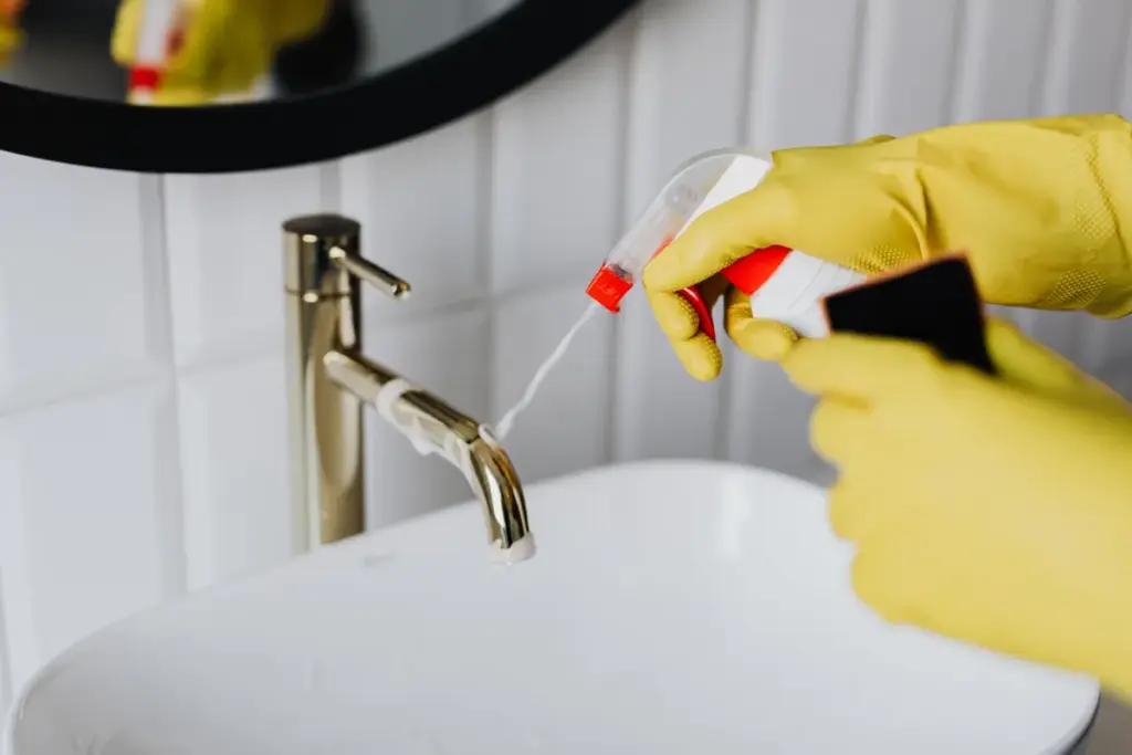 Cleaner in yellow gloves spraying and wiping bathroom sink faucet and mirror with white tiles