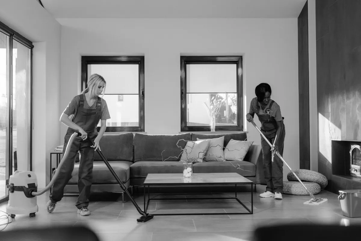 Two cleaners vacuuming a modern open-plan living room with large windows and natural daylight