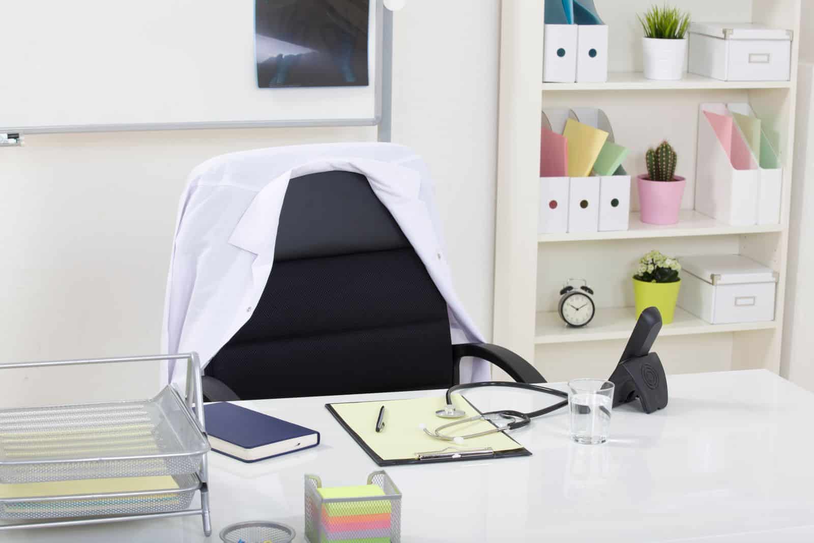 Aufgeräumter Schreibtisch in einem Sprechzimmer mit Stethoskop, Klemmbrett und Regal