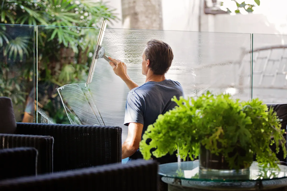 Person cleaning large glass door with squeegee, potted plants on side table visible